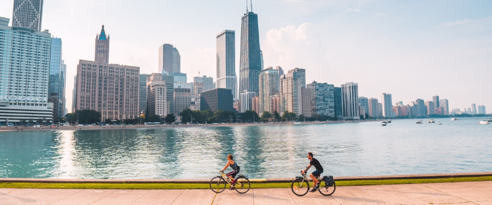 View of the Chicago skyline from Lake Michigan with two people cycling along the promenade in the foreground.
